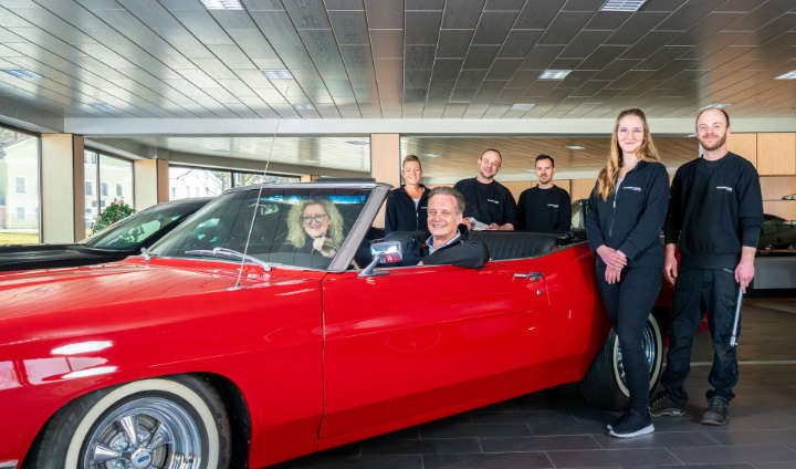 Team eines Autohauses mit rotem Cabrio im Verkaufsraum.